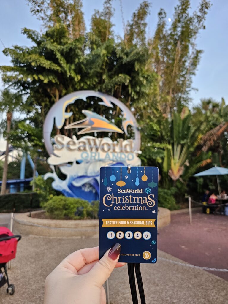  a hand holding out a SeaWorld Christmas Celebration food and drink tasting lanyard in front of the SeawWorld Orlando sign.