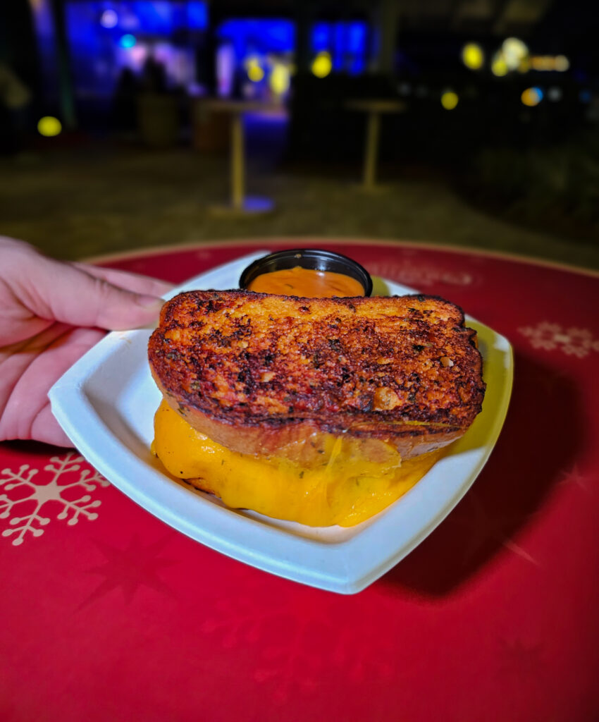a hand holding a plate with a crispy grilled cheese that's oozing out cheese and has a side of tomato soup