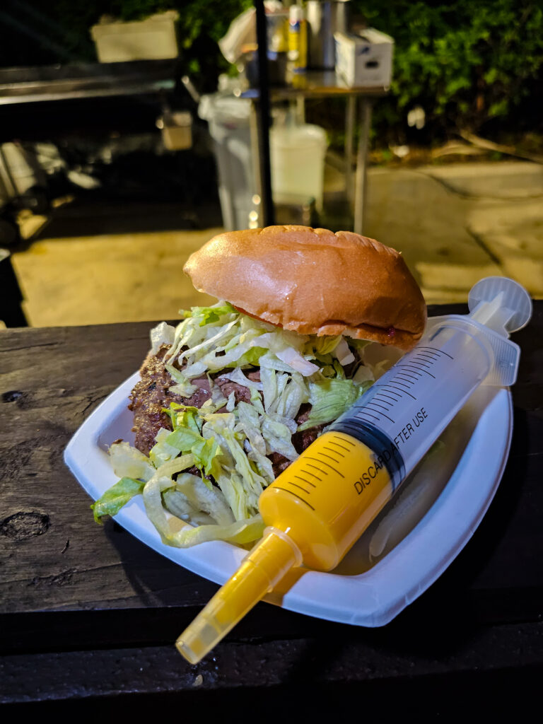 a burger on a plate with the top bun set askew exposing lettuce and a large syringe of cheddar cheese on the side.
